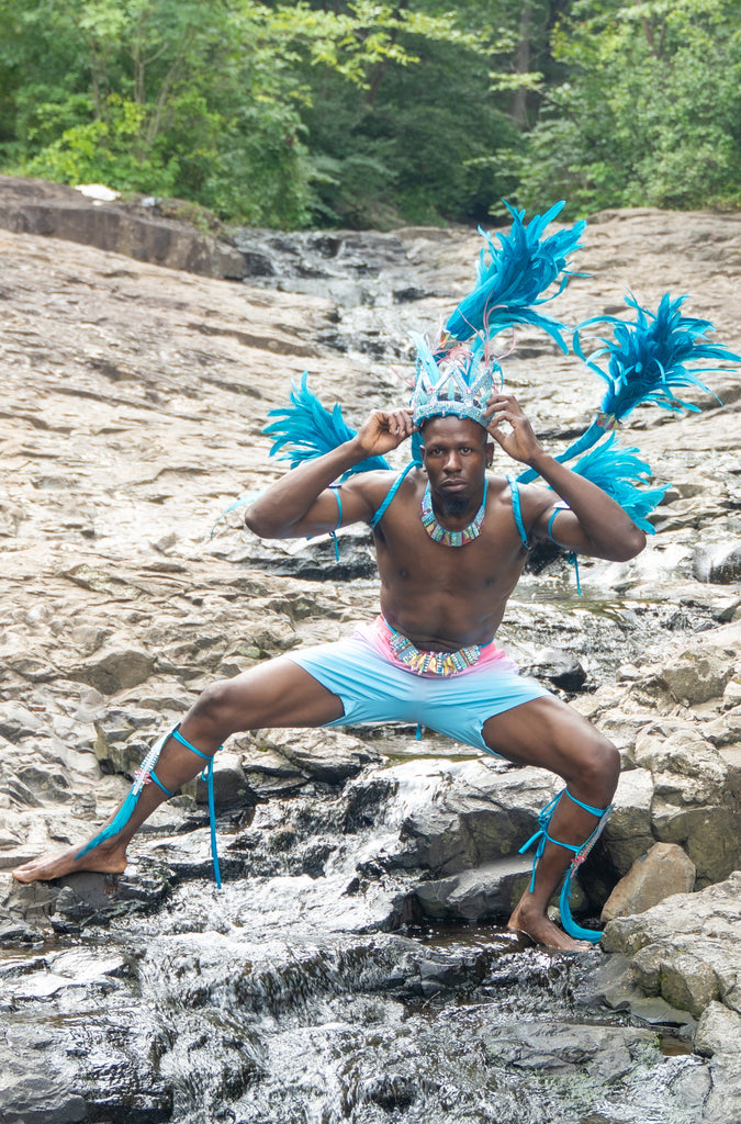 Man in colorful Caribbean costume sitting on a rocky stream with trees in the background. Carnival Costume
Details